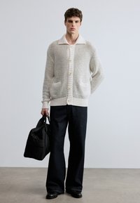 Πλεκτή μπεζ ζακέτα με γιακά, κουμπιά μπροστά και δύο τσέπες, συνδυασμένη με σκούρα τζιν με φαρδύ πόδι και μαύρη τσάντα ώμου.