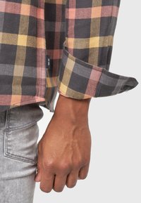 Camisa de franela a cuadros con colores en gris, rosa, amarillo y negro. Mangas con puños y una etiqueta visible con "EST 1993".