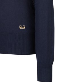 Dunkelblauer Strickpullover mit gerippten Bündchen und Saum. Verfügt über ein goldenes Emblem am unteren linken Rand. Sanfte Textur, klassischer Schnitt.