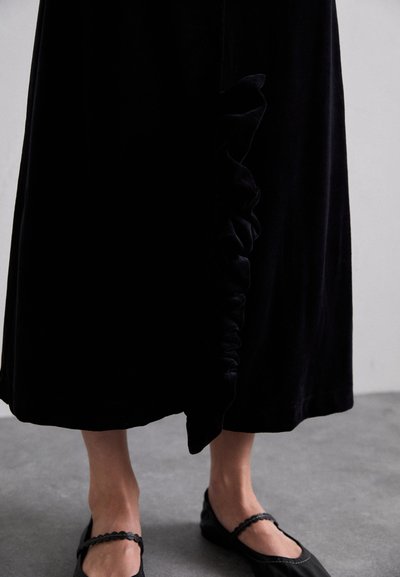 Black velvet maxi dress with side ruffle detail. Paired with black flats featuring a scalloped edge. Smooth texture, elegant silhouette.