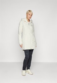 Hvid, fyldig jakke med hætte, der har skrå quiltning og en frontlynlås. Sæt sammen med mørke bukser og lyse sneakers.