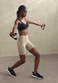 En kvinne i svart sports-BH og beige ribbede shorts bruker treningsbånd med svarte håndtak, iført svarte treningssko på en nøytral gulv.
