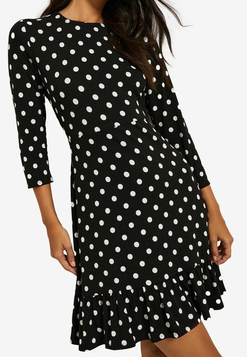 Robe noire longueur genou à pois blancs, manches trois-quarts et ourlet volanté portée par une femme aux cheveux longs et foncés.