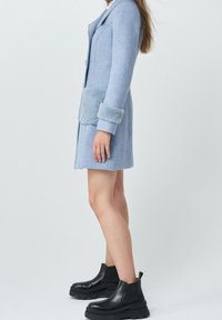 Donna che indossa un cappotto blu chiaro con rifiniture in pelliccia sulle maniche e sulla tasca, abbinato a stivali ankle boots neri e robusti, in piedi di fronte a uno sfondo neutro.