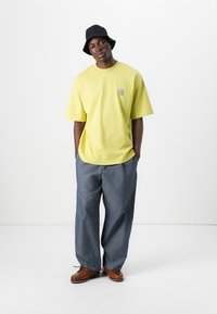 T-shirt jaune taille XL avec un logo sur la poitrine, associé à un pantalon en denim ample et des chaussures à lacets marron. Chapeau seau noir.