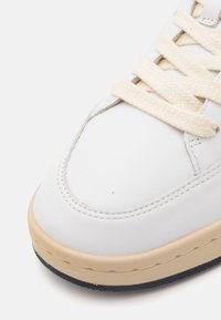 Baskets en cuir blanc avec un bout rond, lacets crème et semelle en caoutchouc beige. Présente des détails de couture et une semelle extérieure texturée bleu marine.