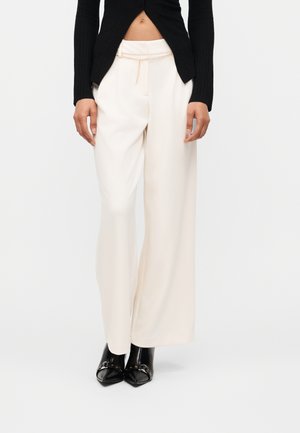 Kobieta nosząca białe spodnie o wysokim stanie i szerokich nogawkach z złotym paskiem w postaci łańcucha, czarny krótki top oraz czarne botki z pointed toe, stojąca prosto.