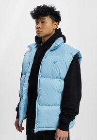 Lysblå puffervest med høj krave, lynlås og store lommer, båret over en sort hættetrøje. Glat tekstur og polstret design.