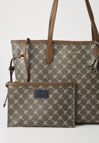 Olivengrønn totebag med hvitt mønster, brune skinndetaljer og en avtagbar matchende pouch med logo. Teksturert overflate.