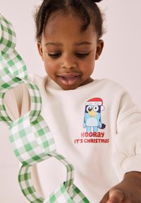Kremowa bluza z krótkim rękawem z niebieskim baśniowym ptakiem i napisem "HOORay IT'S CHRISTMAS." W dłoni zielono-biała papierowa girlanda w kratkę.