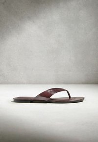 Brun læder thongsandal med flad sål, vist på en lys overflade mod en struktureret grå baggrund.