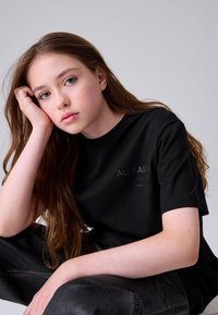 Fiatal, hosszú barna hajú személy fekete ALLSAINTS pólóban és fekete nadrágban, fejét a kezére hajtva, elgondolkodva előre nézve.