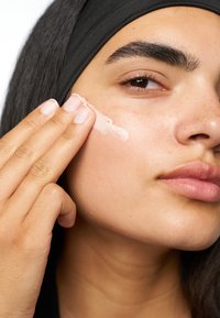 Un primo piano di una persona che applica una crema leggera sulla guancia, mostrando una pelle liscia, un trucco naturale e un elastico per capelli scuro.