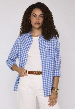 Donna con capelli ricci che indossa una camicia a quadri blu e bianca sopra una canotta bianca e pantaloni con cintura marrone e braccialetti d'oro.