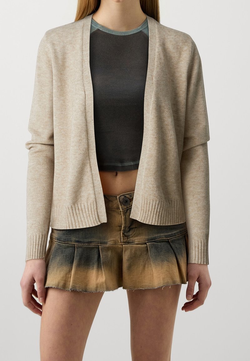 Femme portant un cardigan beige ouvert, un top court gris foncé et une mini-jupe plissée tie-dye marron-noir, debout devant un fond uni.