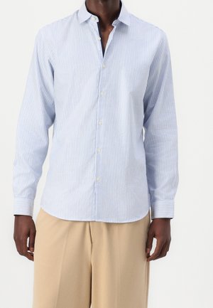 Uomo che indossa una camicia a maniche lunghe a righe verticali blu chiaro e bianca e pantaloni beige larghi, mostrato dalle spalle a metà coscia su uno sfondo bianco.