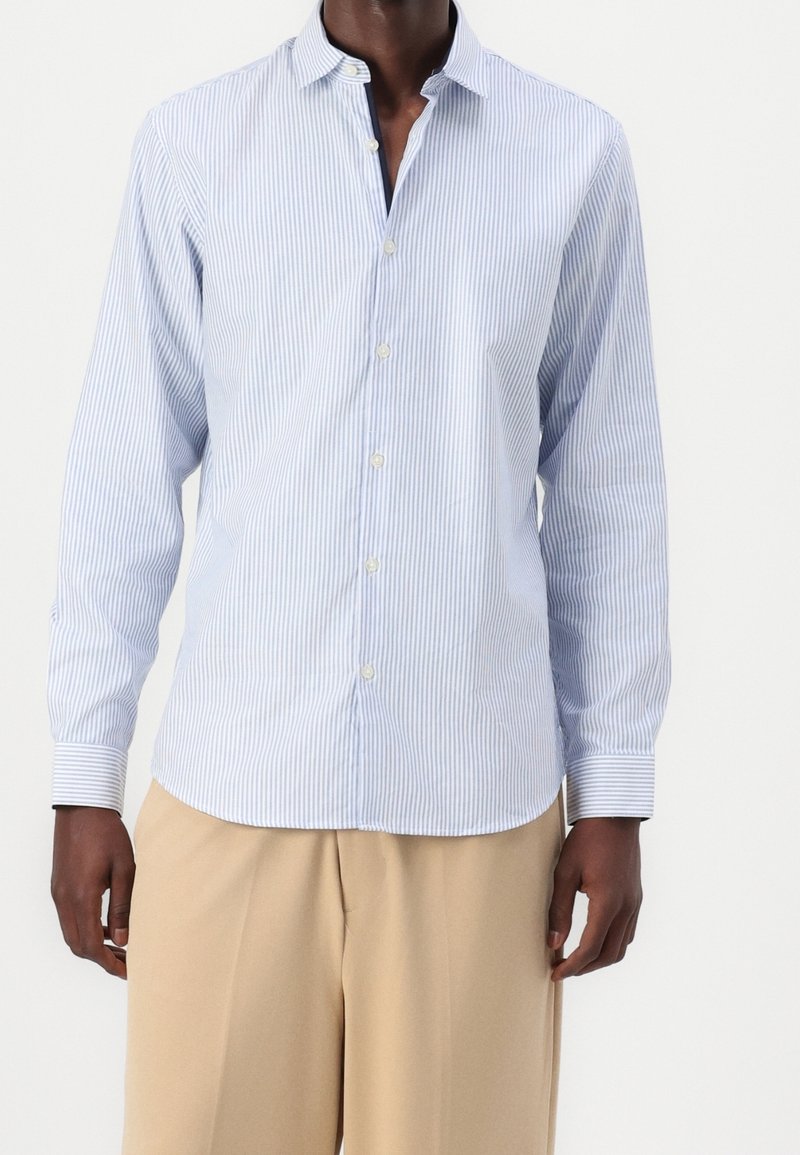 Muž v lehce modré a bílé vertikálně pruhované košili s knoflíky a béžových volných kalhotách, zobrazený od ramen po střed stehna na bílém pozadí.