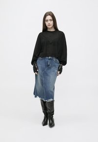 Kvinne iført en svart, gjennomsiktig langermet topp, svarte fingertløse hansker, en slitt denim midi-skjørt og svarte knelang støvler, stående mot en hvit bakgrunn.