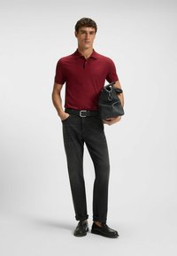 Polo-shirt color bordeaux, jeans neri e scarpe slip-on nere. Porta una borsa nera con texture. Design semplice e forma aderente.