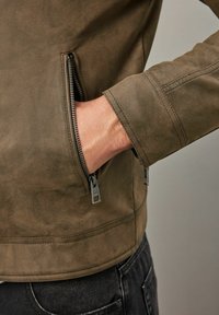 Chaqueta de ante marrón con bolsillos con cremallera y un corte limpio y contemporáneo. Los puños están confeccionados con un acabado suave.