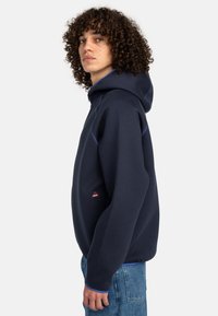 Navy hoodie with a drawstring and light blue stitching details. Features a front logo patch and a relaxed fit. Cozy, stretchy fabric.