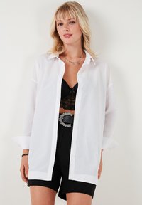 Witte, lichtgewicht button-up shirt met een kraag, lange mouwen en manchetdetails, gelaagd over een zwarte kanttop en gedragen met zwarte shorts.