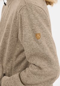 Beige, texturerad huvtröja med dragkedja och ett litet brunt logotypmärke på ärmen. Mjukt tyg med en melerad look.