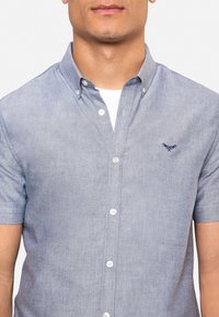 Korte mouw knoop-over-hemd in lichtblauwe chambray stof, met een witte onderlaag, een kraag en een geborduurd logo op de borst.