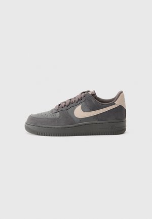 Ciemnoszary zamszowy but sportowy Nike Air Force 1 z beżowym logo i tasiemką na pięcie, płaska podeszwa i sznurówki, widok z boku na jasnoszarym tle.
