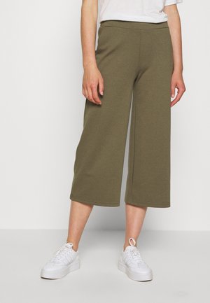 Persoon draagt een olijfgroene, wijd uitlopende cropped broek en witte sneakers, staand tegen een effen witte achtergrond.
