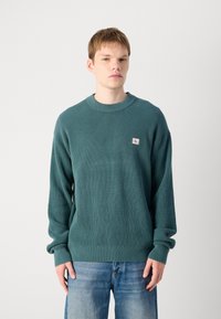 Teal gebreide sweater met een ronde hals, textuur op het oppervlak en geribde manchetten. Bevat een klein logo op de voorkant. Draagt met blauwe jeans.