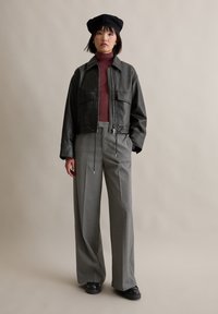 Schwarze,lederne, kurze Jacke mit zwei vorderen Taschen, kastanienbrauner Rollkragenpullover, graue, gestreifte, weite Hosen, schwarze Plateauschuhe und eine schwarze Baskenmütze.