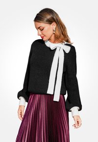 Maglione nero a maglia con un dettaglio a fiocco bianco sul collo; abbinato a una gonna plissettata e lucida di colore bordeaux. I polsini bianchi aggiungono contrasto.