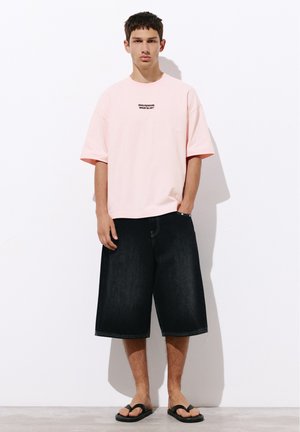 Junger Mann steht und trägt ein hellrosa T-Shirt mit Text, schwarze weite knielange Shorts und schwarze Flip-Flops vor weißem Hintergrund.