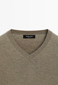Maglione a V in un verde oliva tenui, realizzato in un materiale knit leggero, con rifiniture a costine attorno alcollo e etichetta del brand tessuta.