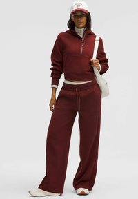 Felpa corta bordeaux con zip a metà, tasca frontale e pantaloni della tuta a gamba ampia coordinati. Accenti bianchi sulla vita e scarpe leggere.