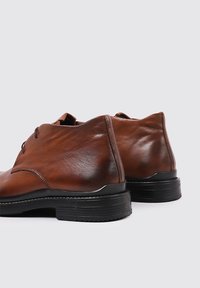 Zapatos de cuero marrón con una textura suave, puntera redondeada, cordones y una suela de goma negra. La altura del tacón es moderada con un detalle mínimo.