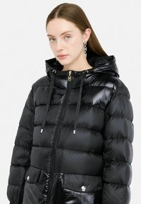 Giacca a vento nera con finitura lucida, chiusura con zip, cordini regolabili e due tasche frontali con dettagli metallici e bottoni.