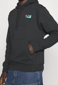 Svart huvtröja med en framficka, med en flerfärgad broderad Nike-logga på bröstet. Tillverkad av mjukt bomullsmaterial.