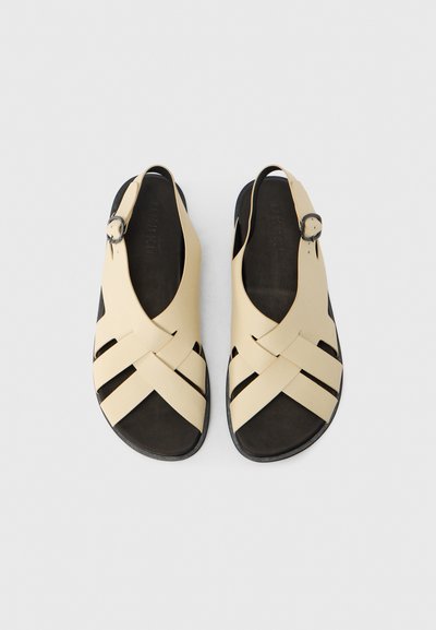 Sandali beige con design a strisce intrecciate, punte arrotondate e una fibbia singola alla caviglia. Plantare nero con texture liscia.