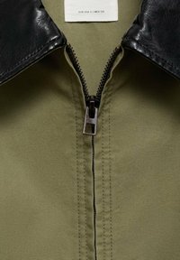 Gros plan d'une veste vert olive avec col en cuir noir et fermeture éclair métallique partiellement fermée, montrant la texture du tissu et les détails des coutures.