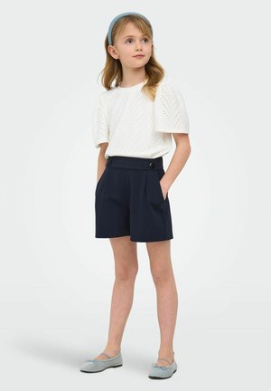 Ragazza giovane che indossa una maglietta bianca a maniche corte con occhielli, pantaloncini blu navy a vita alta con bottoni, ballerine grigie e una fascia per capelli azzurra.