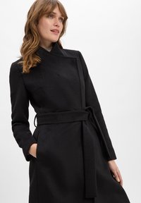 Zwarte jas met een wikkelontwerp, voorzien van een ceintuur in de taille, lange mouwen en een zachte, gestructureerde stof. Inclusief één borstzak.