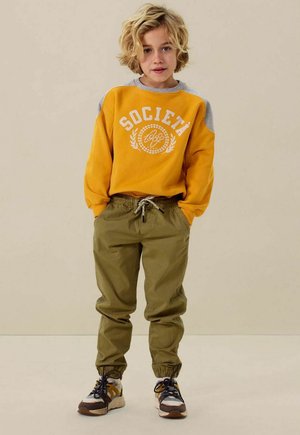 Yellow sweatshirt with gray accents, featuring "SOCIALETÁ 1892" graphic. Olive green jogger pants with elastic cuffs. Sneakers with multi-color details.