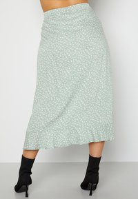 Vrouw in een lichtgroene midi-jurk met witte bloemenprint en gekruiste zoom, gecombineerd met zwarte hoge enkellaarsjes.