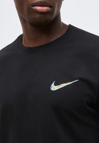 Άνδρας που φοράει μαύρο T-shirt με ιριδίζον λογότυπο Nike swoosh στο αριστερό στήθος, εμφανίζεται από τον λαιμό μέχρι το μέσο του κορμού σε μονόχρωμο φόντο.