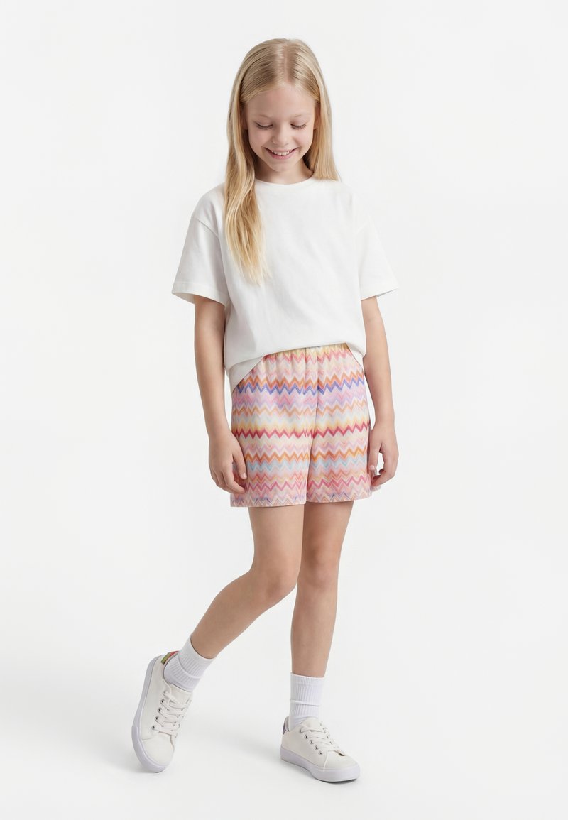 Niña sonriente de pie, con camiseta blanca, pantalones cortos con patrón de zigzag en colores pastel, calcetines blancos y zapatillas blancas sobre un fondo blanco.