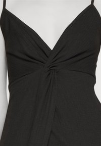 Robe noire sans manches avec fines bretelles et un détail de nœud torsadé au profond décolleté en V sur un buste de mannequin blanc.