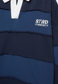 Ragbijska majica z mornarsko modrimi in temno modrimi črtami, belim ovratnikom ter vezenim napisom "STWD COMMUNITY" na prsih.