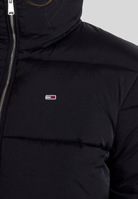 Svart puffjacka med hög krage, prydd med en silverdragkedja och en liten färgglad logotyp på bröstet. Slät yta och quiltad design.